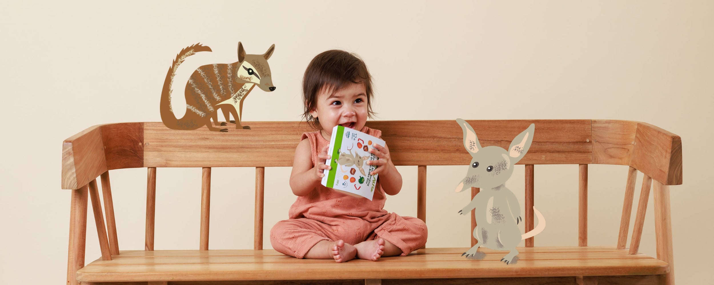 Baby smiling on a bench holding Audrey & Alfie Packet, surrounded by illustrated Australian Animals